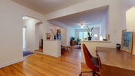 This thoughtfully designed single-floor residence showcases a cohesive layout connected by graceful arched doorways and gleaming hardwood floors throughout. The entry area flows seamlessly into distinct living zones, offering a functional home office nook and a dedicated dining room adorned with a classic chandelier and built-in shelving. The dining area connects through a charming archway to a bright living room featuring a ceiling fan, large windows that invite natural light, and a cozy seating arrangement anchored by a colorful area rug. The galley-style kitchen is well-equipped with warm wood cabinetry, dark countertops, a slate tile backsplash, and black appliances, with a window above the counter providing natural light. The bedroom is a serene retreat with warm hardwood floors, ample storage, and an angled ceiling detail that adds architectural character. The bathroom maintains a classic aesthetic with white subway tile, black accent trim, and a window for natural ventilation. Built-in bookshelves and thoughtful arched passageways throughout the space lend a timeless, refined character, while the open layout and abundant natural light create an airy, welcoming ambiance.