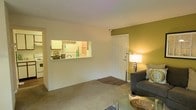 This single-floor residential space offers a functional and welcoming layout designed for comfortable everyday living. The living area is finished with wall-to-wall carpeting that adds warmth underfoot, and features a soothing neutral color palette complemented by a bold olive-green accent wall that anchors the room with character. A stylish floor lamp provides warm ambient lighting, enhancing the inviting atmosphere of the space.

A standout architectural detail is the pass-through window that connects the living area to the kitchen, promoting an open and airy feel while maintaining a distinct separation between the two spaces. The kitchen, visible through this opening, is equipped with white cabinetry and appliances, creating a clean and efficient workspace.

The entry door, finished in crisp white with classic panel detailing, opens directly into the living space, providing a welcoming transition from the exterior. The overall layout is compact yet thoughtfully arranged, making efficient use of the available space. The combination of soft textures, warm lighting, and connected spaces gives this residence a cozy, lived-in quality that is both practical and comfortable.