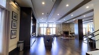 This elegant single-floor space boasts an open-concept design that seamlessly integrates multiple functional areas into one cohesive environment. Rich, dark hardwood flooring runs throughout, anchoring the space with a sense of warmth and refinement. At the heart of the room sits a substantial central island with pendant lighting overhead, ideal for gathering and casual use.

The ceiling features decorative exposed beams with recessed lighting, adding architectural depth and dimension. Large windows along the perimeter flood the interior with generous natural light, creating a bright and welcoming atmosphere. A striking stacked-stone fireplace serves as a dramatic focal point, flanked by a comfortable lounge seating area complete with armchairs and a coffee table arranged on a area rug — perfect for relaxed conversation.

A dedicated media wall with a large flat-screen television is integrated along one side, offering an entertainment-focused zone. The neutral wall tones contrast beautifully with the darker design elements, including the bold accent wall and cabinetry. The overall ambiance is modern yet inviting, blending upscale finishes with practical communal functionality — characteristic of a well-appointed shared amenity or lounge space.