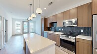 This well-designed one-bedroom, one-bathroom unit features a thoughtfully arranged open-concept floor plan that maximizes its efficient footprint. Light wood-tone flooring runs continuously throughout the space, creating a cohesive and airy feel.

The kitchen serves as a focal point, showcasing flat-front wood-grain cabinetry extending to the ceiling, a striking dark vertical tile backsplash, and a full suite of stainless steel appliances including a range, microwave, dishwasher, and refrigerator. A central island with a white quartz countertop provides additional workspace and seating, illuminated by a trio of modern pendant lights above.

The open living area extends toward large windows and a glass door at the far end, welcoming abundant natural light. The bedroom offers a comfortable, well-proportioned space with dual windows, a ceiling fan, and direct access to the full bathroom. The bathroom features large-format tile, a wood-grain vanity with white countertop, matte black fixtures, and direct entry to a dedicated walk-in closet equipped with wire shelving systems. Clean white walls and modern black hardware accents carry throughout the unit, contributing to its contemporary aesthetic.