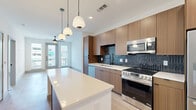 This well-designed one-bedroom, one-bathroom unit features a thoughtfully arranged open-concept floor plan that maximizes its efficient footprint. Light wood-tone flooring runs continuously throughout the space, creating a cohesive and airy feel.

The kitchen serves as a focal point, showcasing flat-front wood-grain cabinetry extending to the ceiling, a striking dark vertical tile backsplash, and a full suite of stainless steel appliances including a range, microwave, dishwasher, and refrigerator. A central island with a white quartz countertop provides additional workspace and seating, illuminated by a trio of modern pendant lights above.

The open living area extends toward large windows and a glass door at the far end, welcoming abundant natural light. The bedroom offers a comfortable, well-proportioned space with dual windows, a ceiling fan, and direct access to the full bathroom. The bathroom features large-format tile, a wood-grain vanity with white countertop, matte black fixtures, and direct entry to a dedicated walk-in closet equipped with wire shelving systems. Clean white walls and modern black hardware accents carry throughout the unit, contributing to its contemporary aesthetic.