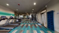 Located at 8718 Wintersweet Ln in Charlotte, NC, this well-appointed fitness center offers a comprehensive workout environment on a single floor. The space is defined by its vibrant, patchwork-style carpet tiles in teal, brown, and neutral tones, adding a modern aesthetic to the functional interior. Recessed lighting throughout provides consistent, even illumination across the entire floor.

The cardio zone features multiple treadmills and an elliptical trainer lined along one wall, each equipped with individual display consoles. A stationary bike and additional cardio machines round out the aerobic options. For strength training, the facility includes a multi-functional cable machine, a dedicated free weights section with a full rack of dumbbells, and a weight bench positioned for versatile use. A barbell and weight plates are also available for compound lifts.

Full-length mirrors span one wall, enhancing the sense of space and allowing users to monitor their form during exercise. A decorative teal stripe accent runs along another wall, tying the color palette together. A BOSU balance trainer and TRX suspension system add functional training options. Natural light filters in through windows on one end, and glass entry doors provide easy access to and from the facility.