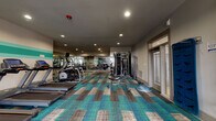 Located at 8718 Wintersweet Ln in Charlotte, NC, this well-appointed fitness center offers a comprehensive workout environment on a single floor. The space is defined by its vibrant, patchwork-style carpet tiles in teal, brown, and neutral tones, adding a modern aesthetic to the functional interior. Recessed lighting throughout provides consistent, even illumination across the entire floor.

The cardio zone features multiple treadmills and an elliptical trainer lined along one wall, each equipped with individual display consoles. A stationary bike and additional cardio machines round out the aerobic options. For strength training, the facility includes a multi-functional cable machine, a dedicated free weights section with a full rack of dumbbells, and a weight bench positioned for versatile use. A barbell and weight plates are also available for compound lifts.

Full-length mirrors span one wall, enhancing the sense of space and allowing users to monitor their form during exercise. A decorative teal stripe accent runs along another wall, tying the color palette together. A BOSU balance trainer and TRX suspension system add functional training options. Natural light filters in through windows on one end, and glass entry doors provide easy access to and from the facility.