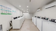 Located in Jacksonville, FL, this shared laundry facility is a practical, well-maintained space designed to serve its users efficiently. The room features a clean, minimalist aesthetic with crisp white walls, white baseboards, and light-toned tile flooring that extends throughout, creating a bright and hygienic environment.

Multiple commercial-grade top-loading washers and dryers are arranged in a row along one wall, providing ample capacity for simultaneous use. Informational charts and usage guides are posted on the walls, offering convenient reference for users. Recessed lighting illuminates the space evenly from above, while a textured ceiling adds a subtle architectural detail.

The facility includes two access points: an interior door and a glass-paneled exterior door that allows natural light to filter in and provides views of the surrounding outdoor greenery. An illuminated exit sign is prominently displayed above the exterior door, ensuring clear emergency egress. A waste bin is conveniently positioned near the exit for easy disposal.

The layout is open and uncluttered, allowing users to move freely between machines. Overall, this laundry facility combines functional design with a clean, well-organized environment, making it a straightforward and accessible amenity for residents at 8220 Dix Ellis Trail.