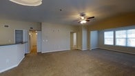 This single-floor space presents an open and airy living area as its central feature. The room is finished with wall-to-wall neutral-toned carpeting that extends throughout, creating a cohesive and comfortable feel underfoot. A ceiling fan with an integrated light fixture is centrally mounted, providing both ambient lighting and air circulation. The walls are painted in a soft, neutral beige tone, complemented by clean white baseboards that frame the space neatly.

Multiple doorways are visible from the main living area, indicating access to additional rooms and spaces beyond. A corridor to the left leads further into the unit, illuminated by overhead lighting and featuring what appears to be a door with vertical paneling. A window on the right side of the space allows natural light to filter in. The layout appears open and functional, with ample floor space that can accommodate a variety of furniture arrangements. A thermostat or control panel is mounted on the wall, adding to the practical features of the space. Overall, the unit offers a clean, move-in-ready environment with a straightforward and efficient floor plan.