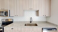 Located at 4500 Swan Ave in St. Louis, MO, this thoughtfully designed single-floor unit offers a clean, contemporary aesthetic throughout. The kitchen serves as a standout feature, showcasing light wood cabinetry with matte black hardware, speckled granite countertops, a white subway tile backsplash, and a full suite of stainless steel appliances including a range, microwave, and refrigerator. An undermount stainless sink with a sleek gooseneck faucet completes the well-appointed cooking space. Wood-look flooring runs throughout the main living areas and bedroom spaces, providing warmth and visual continuity. The hallway connecting the kitchen to the main entrance is bright and open, benefiting from recessed lighting and natural light filtering through slatted entry doors. The bedroom areas feature light-toned flooring, neutral white walls, and ceiling fan fixtures, creating a calm and versatile space. The bathroom is finished with matching light wood cabinetry, granite countertops, a backlit vanity mirror, matte black hardware, and wood-look tile flooring, maintaining the cohesive modern design language found throughout the unit. Overall, the space presents a fresh, neutral palette with modern finishes ideal for comfortable, stylish living.