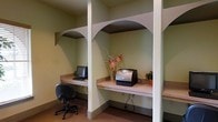 This single-floor business center is designed to provide a functional workspace with a clean, organized layout. The space features a series of built-in workstations separated by structural columns, each framed by distinctive arched details that add a subtle architectural character to the otherwise utilitarian setting. The workstations are equipped with laminate desktop surfaces that offer ample room for computing and printing tasks. A desktop computer and office chair are stationed at one alcove, while an adjacent workstation houses a laser printer, making the area well-suited for a range of business needs. Small decorative touches, including a floral arrangement and a potted plant, add a modest warmth to the space. The walls are painted in a soft sage green, contributing to a calm, neutral ambiance. The carpeted flooring in a warm earth tone complements the overall color palette. The workstations are thoughtfully arranged to provide a degree of separation between users, offering a semi-private environment conducive to focused work.