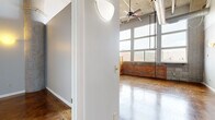 This single-floor loft unit at 1477 Park St in Hartford, CT showcases a striking industrial aesthetic rooted in its original building character. The space is defined by soaring ceilings that amplify the sense of openness throughout, supported by original concrete columns that serve as architectural focal points. Polished concrete floors in rich amber and brown tones extend across the main living areas, reflecting light and adding warmth to the industrial palette.

Large, multi-pane windows dominate one wall, drawing in abundant natural light while framing views of the surrounding rooftops and exposed brick facade. The exposed brick accent wall adds texture and a sense of historic character, complementing the building's converted loft aesthetic. Visible ductwork and ceiling infrastructure reinforce the industrial design language.

The kitchen is outfitted with honey-toned wood cabinetry, dark granite-style countertops, and black appliances including a range, dishwasher, refrigerator, and built-in microwave, all beneath the same dramatic ceiling height. A pendant light anchors the kitchen area.

A separate room with tile flooring provides a functional utility or bathroom space with closet rod storage. The overall ambiance is open, airy, and urban, with a blend of raw industrial elements and practical modern finishes.