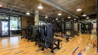 This fitness center showcases a thoughtfully designed single-level layout that combines industrial aesthetics with a fully equipped workout environment. The exposed concrete ceiling with visible ductwork and piping lends a raw, urban character to the space, while pendant-style industrial lights provide focused illumination throughout. The flooring consists of polished hardwood accented with bold colored lane markings, adding both visual interest and functional zoning to the floor plan. A comprehensive collection of black commercial-grade strength machines is arranged throughout the space, including cable machines, leg press stations, and multi-functional training rigs. Large mirrors line portions of the walls, enhancing the sense of depth and allowing users to monitor their form during workouts. Structural concrete columns punctuate the layout, reinforcing the industrial design theme. The generous ceiling height contributes to an open, airy atmosphere despite the density of equipment. The overall ambiance strikes a balance between a professional training facility and a modern, design-forward space, making it both functional and visually engaging for fitness enthusiasts.