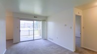 This single-floor space features a welcoming living area finished with wall-to-wall carpeting and clean white walls that lend a bright, neutral aesthetic throughout. A large sliding glass door serves as the focal point of the room, flooding the interior with natural light and providing direct access to a private balcony. The balcony overlooks the surrounding residential community, offering open outdoor views. Vertical blinds accompany the sliding door, allowing flexible control of light and privacy. A floor-level air vent and a wall-mounted thermostat are conveniently positioned, reflecting a practical and functional layout. The ceiling is smooth and painted in a soft, light tone, contributing to the airy feel of the space. A doorway on the adjacent wall leads further into the unit, suggesting additional rooms beyond the main living area. The overall ambiance is clean, simple, and move-in ready, with a neutral palette that accommodates a wide range of interior styles. Natural light, outdoor access, and a straightforward floor plan make this a comfortable and functional living space.