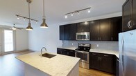 This single-floor space showcases a thoughtfully designed kitchen that serves as a central highlight. Dark espresso-toned cabinetry lines the walls, providing ample storage while creating a bold contrast against the light granite countertops and soft grey subway tile backsplash. Stainless steel appliances, including an electric range and over-the-range microwave, offer a polished, contemporary finish.

A spacious kitchen island anchors the space, featuring the same light granite surface and an undermount sink with a modern arc faucet — ideal for both meal preparation and casual dining. The island's generous proportions make it a functional and social centerpiece.

Lighting plays a key role in the kitchen's ambiance. Industrial-style pendant lights hang above the island, complementing the track lighting system mounted along the ceiling, which provides bright, directed illumination throughout. Light hardwood flooring runs underfoot, warming the overall aesthetic and connecting the kitchen to the adjoining living area.

The open layout promotes a seamless flow between spaces, making this a practical and visually cohesive environment well-suited for everyday living and entertaining.