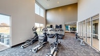 This contemporary fitness center is designed to offer a comprehensive workout experience within a bright, open environment. The main exercise area is anchored by dramatically high ceilings and expansive floor-to-ceiling windows that flood the space with natural light and provide views of the surrounding outdoor landscape. Patterned, multi-tone carpet tiles cover the floors throughout, adding warmth and texture to the modern interior.

The cardio zone is generously appointed with multiple elliptical trainers, treadmills, stationary bikes, and a rowing machine, each positioned to take advantage of the outdoor views and wall-mounted flat-screen televisions. A large overhead ceiling fan ensures comfortable air circulation across the open floor plan.

The strength training area features a well-stocked dumbbell rack displayed in front of full-length mirrors, visually expanding the space and supporting proper form during workouts. Additional cable and weight machines are also available for a complete strength regimen.

A separate studio room, enclosed by sleek floor-to-ceiling glass partition walls and accessible through a glass door, provides a dedicated space for group classes, yoga, or personal training sessions. It features its own windows overlooking the outdoors, hardwood-style flooring, and a ceiling fan.

Completing the facility is a teal-accented nook housing a water fountain and storage shelving, adding both convenience and a pop of color to the overall design.