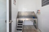 This self-serve dog grooming salon is a purpose-built, single-room space designed with both functionality and cleanliness in mind. The interior is finished with crisp white subway tiles that line the walls from floor to mid-height, creating a hygienic and easy-to-clean environment. Above the tile, smooth white walls carry a bright, open feel throughout the room.

The centerpiece of the space is a large, elevated stainless steel dog wash tub equipped with a coiled blue hose and built-in tethering rails, providing a secure and convenient bathing setup for pets of various sizes. A mounted shelf beside the tub holds grooming essentials, including shampoos and conditioners, readily available for use. A high-powered dryer and broom are also on hand, ensuring a complete grooming experience from wash to dry.

Cheerful, colorful dog-themed artwork is displayed on the upper walls, adding a welcoming and playful touch to the otherwise utilitarian space. A full-length glass exterior door floods the room with natural light and offers a view of the surrounding greenery and parking area outside.

Posted signage outlines usage guidelines and health requirements for visiting pets, reflecting a well-managed and community-oriented facility. Overall, this grooming salon strikes a practical balance between a professional-grade setup and an approachable, pet-friendly atmosphere.