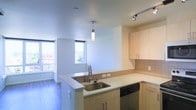 This single-floor space showcases a contemporary open-concept layout, with the kitchen serving as a central design feature. The kitchen is equipped with light wood cabinetry offering generous storage, complemented by a tiled backsplash and light-toned countertops that extend across an L-shaped configuration. A peninsula with an undermount stainless steel sink acts as a natural divider between the kitchen and the adjacent living area, providing both functional workspace and an informal gathering point. Track lighting and a modern pendant light ensure the kitchen is well-illuminated. Stainless steel appliances, including a dishwasher, contribute to the kitchen's clean, modern aesthetic. The living area benefits from multiple large windows that invite abundant natural light and frame views of the surrounding cityscape. Wide-plank hardwood flooring in a cool gray tone runs throughout, lending a cohesive and contemporary feel to the interior. The smooth, light-colored ceilings enhance the sense of openness and airiness. Overall, the space presents a well-designed, functional environment with a clean modern aesthetic and urban appeal.