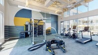 This single-floor amenity facility showcases a thoughtfully designed interior that blends bold aesthetics with practical functionality. The welcoming lobby greets visitors with striking terrazzo-style flooring in mixed tones, soaring ceilings with exposed structural elements, and floor-to-ceiling glass walls that flood the space with natural light. Seating vignettes featuring a green sofa, accent chairs, and a hanging egg chair add character, while sculptural blue pendant lights serve as a dramatic focal point overhead.

Adjacent to the lobby, the leasing and management area features warm wood flooring, a central bar-height counter with teal cabinetry, and a bold chevron-tiled accent wall. A glass-paneled management office provides a semi-private workspace while maintaining visual connectivity to the common area. A cozy nook with a round table, colorful mural wall, and cane-paneled enclosure offers an intimate meeting space.

The fitness center is comprehensively outfitted with treadmills, ellipticals, stationary bikes, a cable machine, battle ropes, free weights, and a wall-mounted storage rack. Large windows offer outdoor courtyard views, and full-length mirrors amplify the sense of space. A vibrant mural with bold geometric shapes in blue, yellow, and green energizes the workout environment throughout.