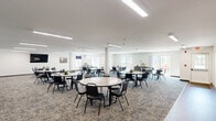Located at 731 E Terra Cotta Ave in Crystal Lake, Illinois, this single-floor multipurpose community room offers a generously sized, open-plan layout well-suited for gatherings, meetings, and communal dining. The main area is carpeted in a neutral patterned tile carpet and is illuminated by recessed linear fluorescent ceiling fixtures that provide consistent, even lighting throughout. Structural columns are integrated into the floor plan, subtly defining zones within the open space without interrupting its overall flow.

The room is furnished with multiple round tables and stackable chairs, accommodating a large number of occupants simultaneously. Along one wall, a built-in kitchenette features dark wood cabinetry, a stainless steel refrigerator, a microwave, and a pass-through window that connects to the main hall — ideal for food service during events.

A wall-mounted flat-screen television is positioned for easy viewing across the room, while a dedicated workstation with a desktop computer offers an additional functional area. The space is equipped with a wall-mounted HVAC unit for climate control.

Sliding glass doors and large windows along one side of the room flood the interior with natural light and provide direct access to an outdoor patio, offering views of the surrounding grounds. The clean, white walls and straightforward design give the space a bright, welcoming, and highly functional character.