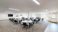 Located at 731 E Terra Cotta Ave in Crystal Lake, Illinois, this single-floor multipurpose community room offers a generously sized, open-plan layout well-suited for gatherings, meetings, and communal dining. The main area is carpeted in a neutral patterned tile carpet and is illuminated by recessed linear fluorescent ceiling fixtures that provide consistent, even lighting throughout. Structural columns are integrated into the floor plan, subtly defining zones within the open space without interrupting its overall flow.

The room is furnished with multiple round tables and stackable chairs, accommodating a large number of occupants simultaneously. Along one wall, a built-in kitchenette features dark wood cabinetry, a stainless steel refrigerator, a microwave, and a pass-through window that connects to the main hall — ideal for food service during events.

A wall-mounted flat-screen television is positioned for easy viewing across the room, while a dedicated workstation with a desktop computer offers an additional functional area. The space is equipped with a wall-mounted HVAC unit for climate control.

Sliding glass doors and large windows along one side of the room flood the interior with natural light and provide direct access to an outdoor patio, offering views of the surrounding grounds. The clean, white walls and straightforward design give the space a bright, welcoming, and highly functional character.
