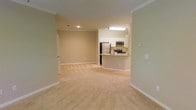 This single-floor space is designed with an open-concept layout that seamlessly connects the living area to the kitchen. The living area features neutral-toned wall-to-wall carpeting that contributes to a warm, comfortable atmosphere throughout the space. Crown molding along the ceiling adds a touch of classic detail, while clean white baseboards provide a crisp contrast to the beige walls.

The kitchen is positioned at the far end of the open floor plan and is outfitted with stainless steel appliances, including a refrigerator, microwave, and range. White cabinetry lines the upper walls, offering ample storage, while a granite-topped breakfast bar serves as both a functional workspace and a natural dividing element between the kitchen and the living area. Recessed and overhead lighting in the kitchen ensures the workspace is well-illuminated.

The overall ambiance of the space is clean, neutral, and move-in ready, with a cohesive color palette of beige and white tones throughout. The open layout enhances the sense of spaciousness, making the area feel airy and versatile for various furniture arrangements.