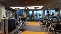 This fitness center is a thoughtfully designed workout facility that offers a comprehensive selection of equipment for both cardio and strength training. The space is defined by its clean, modern aesthetic, featuring a distinctive coffered ceiling with recessed circular lighting fixtures that provide even, focused illumination throughout the room. Large floor-to-ceiling windows line one wall, flooding the interior with natural light and offering open views of the surrounding exterior — creating an energizing atmosphere for exercise. The flooring is a combination of supportive carpet-style material and bold orange rubber matting, delineating workout zones while providing comfort and safety underfoot. A row of treadmills is positioned along the windowed wall, allowing users to enjoy the natural light and views during cardio sessions. Elliptical trainers and stair climbers are arranged nearby, rounding out the cardio section. The strength training area features a range of cable machines and resistance equipment, including Matrix-branded units, offering versatile options for full-body workouts. The layout is open and spacious, allowing for easy movement between equipment. Wall-mounted mirrors enhance the sense of space. Overall, this fitness center combines functionality with a polished, welcoming environment.
