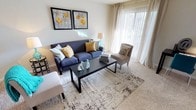 This single-floor model space showcases a thoughtfully designed living area that balances comfort and style. The living room is anchored by plush, neutral-toned wall-to-wall carpeting that adds warmth underfoot, while a large sliding glass door dressed in sheer white vertical blinds allows generous natural light to flood the interior, creating an airy and inviting atmosphere.

The space is furnished with a navy blue sofa accented by a mix of teal, yellow, and patterned throw pillows, complemented by a cushioned accent chair in a soft gray tone. A sleek, dark rectangular coffee table sits atop a patterned area rug, grounding the seating arrangement and adding visual depth to the room. Decorative elements, including a pair of framed floral wall prints in yellow and gray, a teal table lamp, and ceramic bird figurines, contribute to a cohesive and curated aesthetic.

The neutral wall tones provide a versatile backdrop that enhances the brightness of the space, while the open layout promotes a sense of ease and flow throughout. Overall, the living area presents a clean, modern design well-suited for comfortable everyday living.