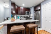 Located at 20 Terrace Avenue in Hasbrouck Heights, NJ, this single-floor residence offers a well-organized and functional layout. The kitchen is a standout feature, outfitted with rich dark espresso cabinetry, quartz countertops, and a full suite of stainless steel appliances including a gas range, built-in microwave, dishwasher, and refrigerator. A classic white subway tile backsplash adds a clean, timeless aesthetic, while a peninsula with seating provides additional workspace and casual dining. The living area is warm and inviting, featuring classic hardwood flooring throughout. Natural light filters in through windows dressed with layered black and sheer curtains, and a glass coffee table anchors the space. The entry door opens directly into this room, which also features a wall-mounted air conditioning unit and a traditional radiator for year-round comfort. The bedroom is a quiet retreat with the same hardwood flooring, offering ample space for a large bed and storage furniture. A window with dual-layer curtains allows for flexible light control. The overall aesthetic throughout the home is cohesive, with cool grey walls providing a neutral, modern backdrop that ties each room together seamlessly.