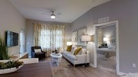 This single-floor residence offers a well-appointed and cohesive living environment designed for both comfort and style. The living area serves as the centerpiece of the space, showcasing vaulted ceilings that lend an open, airy feel to the room. A ceiling fan with integrated lighting anchors the space overhead, while warm hardwood-style flooring runs throughout, adding a touch of natural warmth underfoot. Large windows dressed in patterned gold and white curtains flood the room with abundant natural light, creating a bright and welcoming atmosphere. The living space is elegantly furnished with a neutral-toned sofa accented by gold and patterned throw pillows, complemented by a sleek round white coffee table and a dark leather armchair. Thoughtful lighting, including a tall floor lamp and coordinating table lamps, adds layers of warmth throughout. Abstract artwork adorns the gray-toned walls, enhancing the modern aesthetic. An open passageway connects the living area to the bedroom, which is visible in the background and continues the space's calm, neutral palette with soft bedding and subtle lamp lighting. The overall ambiance is refined, relaxed, and move-in ready.