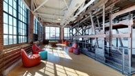 Roebling Lofts is a remarkable single-floor space that masterfully blends industrial heritage with modern functionality. The expansive open layout is defined by soaring white-painted truss ceilings that draw the eye upward, flooding the space with an airy, open atmosphere. Original exposed brick walls line the perimeter, while massive steel cross-braced structural columns and riveted ironwork serve as striking architectural focal points, preserving the building's storied industrial past. Large multi-pane factory-style windows line the walls, inviting abundant natural light that casts dramatic patterns across the warm hardwood floors throughout the day. The interior is thoughtfully appointed with vibrant, modern furnishings — including colorful lounge chairs and ottomans — that create a dynamic contrast against the raw, historic materials. A billiard table anchors a central recreation area, adding to the space's versatile, communal character. The interplay between original industrial elements and contemporary design details gives Roebling Lofts a unique, energetic ambiance that is both visually compelling and highly functional, making it an exceptional destination that honors its architectural roots while embracing modern living.