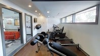 This single-floor fitness room offers a focused and functional workout environment designed to accommodate a variety of exercise routines. The space features warm wood-style flooring that contrasts cleanly against crisp white walls, creating a bright and motivating atmosphere. Recessed lighting is evenly distributed across the dark-trimmed ceiling, providing consistent illumination throughout the room.

The gym is equipped with multiple cardio machines, including stationary bikes and what appears to be a rowing or resistance bench in the foreground, allowing for both low-impact and high-intensity training options. A wall-mounted flat-screen television is positioned for easy viewing during workouts, while wall-mounted exercise balls add both functional storage and decorative appeal. The equipment is arranged to maximize the available floor space without feeling cluttered.

Natural light filters in through horizontal windows positioned along the upper portion of one wall, offering a glimpse of outdoor greenery and contributing to the open feel of the space. A partially visible adjacent room or hallway suggests additional amenity space nearby. Overall, the fitness room is thoughtfully laid out to deliver a comprehensive exercise experience in a clean, modern setting.