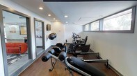 This single-floor fitness room offers a focused and functional workout environment designed to accommodate a variety of exercise routines. The space features warm wood-style flooring that contrasts cleanly against crisp white walls, creating a bright and motivating atmosphere. Recessed lighting is evenly distributed across the dark-trimmed ceiling, providing consistent illumination throughout the room.

The gym is equipped with multiple cardio machines, including stationary bikes and what appears to be a rowing or resistance bench in the foreground, allowing for both low-impact and high-intensity training options. A wall-mounted flat-screen television is positioned for easy viewing during workouts, while wall-mounted exercise balls add both functional storage and decorative appeal. The equipment is arranged to maximize the available floor space without feeling cluttered.

Natural light filters in through horizontal windows positioned along the upper portion of one wall, offering a glimpse of outdoor greenery and contributing to the open feel of the space. A partially visible adjacent room or hallway suggests additional amenity space nearby. Overall, the fitness room is thoughtfully laid out to deliver a comprehensive exercise experience in a clean, modern setting.