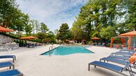This resort-style pool area offers a well-appointed outdoor amenity space designed for relaxation and leisure. At the center of the space is a generously sized swimming pool with a classic geometric layout, trimmed with blue tile accents and clearly marked depth indicators, making it suitable for a range of swimmers. The surrounding pool deck is finished in a light-toned concrete surface that provides ample space for lounging.

Scattered throughout the deck are modern blue chaise lounge chairs paired with vibrant orange umbrellas, offering shaded retreat options at multiple points around the pool. A standout feature of this space is the cluster of hammocks suspended between wooden posts on the pool's edge, adding a relaxed, resort-inspired atmosphere. Additional seating areas with patio furniture are tucked along a raised stone retaining wall, which adds a natural, textured architectural element and creates a tiered visual effect.

The entire area is enveloped by mature trees — including tall pines and broad magnolias — providing a lush green canopy that enhances the sense of privacy and seclusion. A stone accent wall at the upper level, along with a pergola-style structure, adds architectural interest and defines separate zones within the space. The combination of natural landscaping and thoughtfully arranged amenities creates a tranquil, vacation-like atmosphere throughout.