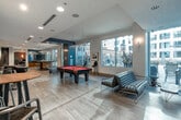 This expansive single-floor amenity space is designed to offer a well-rounded mix of recreation, relaxation, and productivity. The layout flows through several distinct zones, each with its own character and function, all unified by light wood-tone flooring, recessed lighting, and a contemporary design palette of blues, grays, and warm wood accents.

The main lounge and game area anchors the space, featuring a full-size pool table with a bold red felt surface, sleek leather swivel chairs, and open sightlines that create an inviting, social atmosphere. Large windows along the perimeter allow generous natural light and views of the surrounding urban streetscape. Adjacent seating areas offer tufted green sofas, modern side tables, and curated greenery that add warmth and texture.

A dedicated screening room provides a more intimate retreat, outfitted with classic leather lounge chairs and ottomans, a wall-mounted flat-screen television, and a coffered ceiling with recessed lighting for an enhanced viewing experience.

The business center includes a built-in desk with a desktop computer, printer, and pendant lighting, accompanied by seating for quiet work or reading. A compact kitchenette with wood cabinetry, dark countertops, a sink, and a coffee station supports everyday convenience throughout the space.