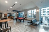This expansive single-floor amenity space is designed to offer a well-rounded mix of recreation, relaxation, and productivity. The layout flows through several distinct zones, each with its own character and function, all unified by light wood-tone flooring, recessed lighting, and a contemporary design palette of blues, grays, and warm wood accents.

The main lounge and game area anchors the space, featuring a full-size pool table with a bold red felt surface, sleek leather swivel chairs, and open sightlines that create an inviting, social atmosphere. Large windows along the perimeter allow generous natural light and views of the surrounding urban streetscape. Adjacent seating areas offer tufted green sofas, modern side tables, and curated greenery that add warmth and texture.

A dedicated screening room provides a more intimate retreat, outfitted with classic leather lounge chairs and ottomans, a wall-mounted flat-screen television, and a coffered ceiling with recessed lighting for an enhanced viewing experience.

The business center includes a built-in desk with a desktop computer, printer, and pendant lighting, accompanied by seating for quiet work or reading. A compact kitchenette with wood cabinetry, dark countertops, a sink, and a coffee station supports everyday convenience throughout the space.