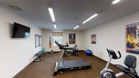 Located at 659 Clough Pike in Cincinnati, Ohio, this fitness center offers a well-equipped, single-floor space designed to support a well-rounded workout routine. The room is illuminated by bright overhead fluorescent strip lighting, creating a clean and energizing atmosphere throughout. Durable, dark-toned carpet flooring covers the entire space, providing comfort and stability underfoot.

The cardio section features a treadmill, a Schwinn upright exercise bike, a recumbent bike, and additional cardio equipment, offering a variety of options for aerobic training. A multi-function cable weight machine anchors the strength-training area, complemented by a flat weight bench and a wall-mounted dumbbell rack stocked with free weights in various sizes. A stability ball is also available for functional fitness exercises.

A wall-mounted flat-screen television provides entertainment during workouts, while framed dumbbell exercise reference charts displayed on the walls serve as helpful guides. Vintage-style athletic posters add a touch of personality to the otherwise clean, neutral-toned walls. A window along one wall allows for a degree of natural light. The space is accessed through a secure door with a glass panel, and a fire alarm system is visibly installed, ensuring safety throughout the facility.