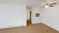 Located at 5782 Andrews Rd. in Mentor on the Lake, Ohio, this single-floor unit offers a straightforward and functional layout designed for comfortable everyday living. The main living area is generously proportioned, featuring wall-to-wall carpeting that adds warmth underfoot and crisp white walls that keep the space feeling bright and clean. A ceiling fan with a light fixture provides both ventilation and ambient lighting to the living space. A baseboard heating system runs along the perimeter walls, ensuring efficient climate control throughout the cooler months. A defining architectural detail is the pass-through window connecting the living area to the kitchen, allowing for open sightlines and easy interaction between spaces. The kitchen area features red-toned cabinetry and includes a refrigerator, with additional appliances visible in the utility area nearby. A hallway connects the main living space to the remaining rooms, with a louvered closet door visible along the corridor, providing practical storage. Natural light filters into the living areas through windows positioned along the exterior walls, contributing to a welcoming interior atmosphere. The overall layout is compact yet efficient, with each area clearly defined and easy to navigate, making this a practical and livable single-story space.