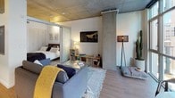 This single-floor unit showcases a thoughtfully designed open-concept layout that seamlessly integrates the living, kitchen, and sleeping areas. The exposed concrete ceiling is a defining architectural feature, lending an industrial character that is balanced by warm light wood flooring running throughout the space. Structural concrete columns further reinforce the loft-inspired aesthetic.

The kitchen is fitted with sleek white cabinetry, a subway tile backsplash, stainless steel appliances including a dishwasher, range, and refrigerator, and light countertops that create a clean, modern look. The kitchen flows directly into the living area, which benefits from abundant natural light provided by expansive floor-to-ceiling windows. These windows offer views of the surrounding urban environment and open to a small outdoor area.

The bedroom is separated from the living space by a sliding glass door, offering privacy while maintaining the open feel of the unit. The bathroom features a white undermount sink set in a modern vanity with clean-lined cabinetry, a large mirror, chrome fixtures, and a shower with a textured tile accent wall, all complemented by wood-look flooring.