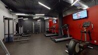 This fitness facility offers a comprehensive workout environment spread across a single floor, designed with an industrial-modern aesthetic that combines exposed ductwork, corrugated metal ceilings, and bold red accent walls against a neutral grey and white palette. Structural columns add a distinctive architectural character throughout the space.

The facility is divided into two primary zones. The strength and cardio area features rubber flooring for safety and durability, housing a well-rounded selection of equipment including treadmills, ellipticals, cable machines, resistance equipment, a dumbbell rack, and benches. Wall-mounted flat-screen televisions are positioned above the cardio machines for entertainment during workouts. This zone is illuminated by long linear fluorescent fixtures that provide bright, even lighting throughout.

The multipurpose studio area is finished with smooth hardwood-style flooring, offering a versatile open layout ideal for group classes, cycling sessions, and functional training. It features a row of spin bikes, a freestanding training dummy, stability balls, resistance bands, and additional accessories neatly organized on storage racks. Full-length mirrors with red trim line one wall, and large windows allow natural light to fill the room. The overall ambiance is energetic and purposeful, making it a functional space suited for a wide variety of fitness activities.