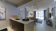 This thoughtfully designed single-floor residence showcases a seamless open-concept layout that connects the kitchen, dining, and living areas into one cohesive and functional living space. The contemporary kitchen features dark wood cabinetry with clean lines, a light-toned countertop island with an undermount sink, a professional-style pull-down faucet, and track lighting overhead that highlights the space with a modern industrial flair. The island serves as both a prep space and a natural divider between the kitchen and the adjoining dining and living areas. The dining area is anchored by a wooden table surrounded by mid-century modern style chairs, complemented by a stylish pendant light above. A vivid yellow accent wall and curated artwork add warmth and personality to the space. The living area is bathed in abundant natural light streaming through expansive floor-to-ceiling windows, offering a bright and airy atmosphere. A ceiling fan adds comfort overhead. Light hardwood-style flooring flows throughout, tying together the clean, contemporary aesthetic. The overall ambiance is modern, vibrant, and inviting, ideal for both everyday living and entertaining.
