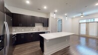 This single-floor unit presents a contemporary design with a cohesive aesthetic throughout. The kitchen serves as a central highlight, featuring dark espresso shaker-style cabinetry that extends to the ceiling, contrasted by crisp white quartz countertops and a matching oversized island. A textured stone tile backsplash adds visual depth, while stainless steel appliances — including a French door refrigerator, range, microwave, and dishwasher — provide a polished, functional finish. Stylish pendant lighting hangs above the island, complementing the recessed lighting overhead. Rich dark hardwood-style flooring flows through the main living and kitchen areas, creating a warm, grounded atmosphere.

The bedrooms are finished with neutral carpet and receive natural light through windows fitted with blinds. One bedroom features French doors, enhancing its connection to natural light. The primary bathroom is generously proportioned, offering a dual-sink dark vanity with white countertops, a soaking tub, tiled shower, light tile flooring, and access to a spacious walk-in closet with built-in shelving. A secondary bathroom mirrors the design language with a single dark vanity, white countertop, and coordinating tile flooring.