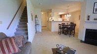 This two-story residence offers a thoughtfully designed open-concept main level that integrates the living, dining, and kitchen areas into a cohesive and functional space. The living area features plush carpeting underfoot, creating a comfortable and inviting atmosphere, and is anchored by a sofa that faces toward the rest of the main floor.

The dining area is positioned adjacent to the kitchen and features a raised bar-height table with matching dark wood chairs, illuminated overhead by an elegant chandelier with warm-toned lighting. This seating arrangement creates a natural gathering point between the living space and the kitchen.

The kitchen is defined by a raised peninsula that provides both a visual and functional divide from the dining area. White cabinetry lines the upper walls, complemented by warm wood-toned lower cabinets, and overhead pendant lighting adds a finishing touch. A front entryway is visible in the background, featuring a tiled floor that transitions from the carpeted main living space.

A carpeted staircase along the left side of the main level leads to the second floor, adding vertical dimension to the layout. The overall ambiance is light and neutral, with soft tones throughout contributing to a clean, understated aesthetic.