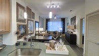 This single-floor space showcases a thoughtfully designed open-concept layout that seamlessly connects the kitchen and living areas, creating a cohesive and inviting atmosphere. The kitchen features a sleek countertop with a contemporary undermount sink and modern pendant lighting overhead, providing both functionality and visual appeal. A curated display of framed artwork lines the walls near the kitchen, adding a gallery-like sophistication to the space. The living area is anchored by a comfortable sofa and complementary seating arranged around a low-profile coffee table, ideal for both relaxed lounging and entertaining. A ceiling fan with a brushed metal finish is centrally positioned to enhance airflow and comfort. Large windows dressed with horizontal blinds and dark drape panels allow natural light to fill the room while offering flexible privacy control. A dark wood dresser or credenza along one wall adds storage and contrast to the lighter tones of the space. Track and pendant lighting work in concert to create warm, layered illumination throughout. The overall ambiance is polished and contemporary, with a neutral color palette punctuated by bold decorative accents, framed prints, and carefully styled surfaces that reflect an elevated, design-forward sensibility.