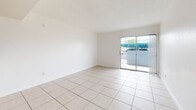 Located at 1515 W Missouri Ave in Phoenix, AZ, this single-floor, one-bedroom, two-bathroom unit offers a functional and straightforward layout with consistent light-toned tile flooring throughout. The open living area is bright and airy, featuring a textured ceiling, clean white walls, and a sliding glass door that invites natural light and provides direct access to an outdoor patio area. The kitchen connects to the living room via a pass-through opening, offering white cabinetry, grey laminate countertops, a built-in sink, and a tiled backsplash accent, making efficient use of the available space. A hallway leads from the main living area to the private bedroom, which benefits from natural light through a window fitted with vertical blinds. Built-in shelving is accessible from the hallway, providing additional storage. Both bathrooms are finished with tile flooring; one includes a full bathtub and shower combination with tiled surround walls and a grab bar. The overall design is clean and minimal, with white walls and neutral finishes throughout, creating a bright, low-maintenance living environment suited for practical everyday use.