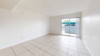 Located at 1515 W Missouri Ave in Phoenix, AZ, this single-story unit offers a practical and efficient layout with one bedroom, two bathrooms, and a combined kitchen and living area. Light-colored ceramic tile flooring runs consistently throughout the entire unit, creating a cohesive and easy-to-maintain interior.

The open-concept living room is bright and airy, with white walls, a textured ceiling, and a sliding glass door that fills the space with natural light while providing access to an outdoor area with views of the surroundings. A pass-through opening connects the living room to the kitchen, enhancing the sense of openness. The kitchen features white cabinetry, gray laminate countertops, a tile backsplash accent, and a full-size refrigerator.

A hallway leads from the main living area to the bedroom and bathrooms. The bedroom offers a comfortable footprint with a window that brings in additional natural light. Both bathrooms are finished with tile flooring, and at least one includes a full tub and shower combination with tiled surround walls. A built-in shelving unit is visible in the hallway, offering convenient storage. The unit's clean, white interior palette throughout gives the space a bright and functional feel.