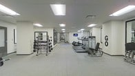 This fitness center offers a comprehensive workout environment designed to accommodate a wide range of exercise routines. The space is laid out on a single floor with a long, open floor plan that provides ample room for movement and equipment placement. The flooring consists of durable, textured fitness tiles that offer both safety and comfort during workouts. Overhead, recessed panel lighting illuminates the entire space evenly, creating a bright and energizing atmosphere with a clean white ceiling and walls throughout. Along one side of the room, a rack of dumbbells in graduated weights is positioned in front of a large wall mirror, supporting free weight and strength training exercises. On the opposite side, a row of treadmills and additional cardio machines are lined up, offering dedicated space for cardiovascular workouts. A wall-mounted flat-screen television is positioned for easy viewing during exercise. Further into the space, additional strength and resistance training equipment is visible, making use of the full depth of the room. The overall ambiance is clean, functional, and professional, reflecting a well-maintained facility suited for daily fitness use.