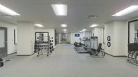 This fitness center offers a comprehensive workout environment designed to accommodate a wide range of exercise routines. The space is laid out on a single floor with a long, open floor plan that provides ample room for movement and equipment placement. The flooring consists of durable, textured fitness tiles that offer both safety and comfort during workouts. Overhead, recessed panel lighting illuminates the entire space evenly, creating a bright and energizing atmosphere with a clean white ceiling and walls throughout. Along one side of the room, a rack of dumbbells in graduated weights is positioned in front of a large wall mirror, supporting free weight and strength training exercises. On the opposite side, a row of treadmills and additional cardio machines are lined up, offering dedicated space for cardiovascular workouts. A wall-mounted flat-screen television is positioned for easy viewing during exercise. Further into the space, additional strength and resistance training equipment is visible, making use of the full depth of the room. The overall ambiance is clean, functional, and professional, reflecting a well-maintained facility suited for daily fitness use.