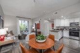 Located at 2200 Colorado Avenue in Santa Monica, CA, this well-appointed one-bedroom, one-bathroom residence offers a thoughtfully designed layout across a single floor. The open-concept living, dining, and kitchen area serves as the heart of the home, bathed in natural light and anchored by warm wood-style flooring. The living room features neutral tones, track lighting, and a sliding glass door that opens onto a private balcony with lush greenery views. The kitchen is outfitted with white shaker cabinetry, stainless steel appliances including a range, microwave, dishwasher, and refrigerator, complemented by grey countertops and recessed lighting. The dining area accommodates a round table with ample space, positioned alongside a closet and a large wall mirror that reflects the kitchen beyond. The bedroom offers a serene retreat with a window framing outdoor foliage, a distinctive geometric accent wall, and a dedicated walk-in closet with built-in shelving. The full bathroom includes a vanity with under-cabinet storage, built-in wall shelving, and a full tub-shower combination. Wood-style flooring and a cohesive neutral palette unify the space throughout.