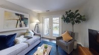 This single-floor residence offers a thoughtfully designed layout that balances comfort and functionality. The open-concept living and dining area serves as the heart of the home, featuring dark hardwood-style flooring, neutral gray walls, and a set of French doors that flood the space with natural light while providing direct access to an outdoor patio. The living area is generously proportioned, offering ample room for seating arrangements centered around a media console.

The kitchen is well-equipped with stainless steel appliances — including a full-size refrigerator, electric range, and built-in microwave — complemented by crisp white cabinetry extending to the ceiling for abundant storage. Dark granite countertops and a stainless steel sink complete the polished, modern aesthetic. Wood-style flooring continues throughout the kitchen and dining area, creating a cohesive flow.

The primary bedroom is a comfortable retreat with plush carpeting, soft neutral tones, and a large window offering views of lush greenery outside. A white dresser and mirror add a clean, bright accent to the room. A second bedroom provides comparable comfort, also featuring carpet, a window with natural light, and a reach-in closet with built-in shelving for organized storage.

The bathroom features a full tub and shower combination with chrome fixtures and a decorative floral shower curtain, maintaining the space's clean and inviting aesthetic throughout.
