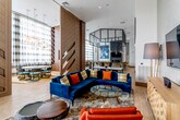 This elegant single-floor amenity lounge showcases a bold contemporary design with soaring double-height ceilings that lend an airy, expansive feel to the entire space. Large floor-to-ceiling windows dressed with sheer plaid curtains flood the interior with abundant natural light, while warm-toned tile flooring flows seamlessly throughout.

The fully equipped kitchen and bar area serves as a central hub, featuring dark granite countertops, a large island, stainless steel appliances, and yellow upholstered barstools that add a vibrant pop of color. Sleek cone-shaped pendant lights hang dramatically above, complemented by an industrial-style black steel transom window frame that divides the upper wall.

A series of richly paneled wooden dividers with geometric chevron detailing define distinct zones within the open-plan layout, including an intimate dining area with a round table and stylish chairs, and a relaxed lounge zone anchored by a deep navy sectional sofa adorned with bold orange and mustard accent cushions. A wall-mounted flat-screen TV and a herringbone-patterned credenza complete the media corner.

A cozy window nook with built-in upholstered bench seating offers a quiet retreat, adorned with curated artwork and accent pillows. Throughout the space, carefully selected art pieces and a warm, curated color palette of navy, amber, orange, and walnut tones create a refined yet inviting communal atmosphere.