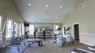 Located at 5303 Wilcox Rd in Dublin, Ohio, this single-floor fitness center is designed to support a well-rounded exercise routine in a bright, welcoming environment. The space features soaring vaulted ceilings that create an open, airy atmosphere, complemented by recessed lighting and natural light streaming in through multiple windows. A full-length wall mirror spans one side of the room, visually expanding the space and allowing users to monitor their form during workouts. The facility is equipped with a selection of cardio machines, including elliptical trainers, as well as a comprehensive free weight station featuring a full dumbbell rack. Strength training is further supported by adjustable benches and additional resistance equipment. A dedicated stretching and floor exercise area with mat space is also available. A wall-mounted television adds a convenient entertainment option during workouts. Neutral-toned walls, carpeted flooring, and a ceiling fan contribute to a comfortable and functional training environment, making this fitness center a practical and well-appointed amenity for its users.