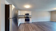 This single-floor residence offers a clean, modern aesthetic with thoughtfully chosen finishes throughout. The open-concept kitchen flows seamlessly into the living area, creating a spacious and functional layout. The kitchen is equipped with white shaker-style cabinetry, granite-look countertops, and a full suite of stainless steel appliances, including a refrigerator, dishwasher, electric range, and built-in microwave. Light wood-tone luxury vinyl plank flooring runs through the kitchen, living, and entry areas, lending warmth and easy maintenance to the main living spaces. The living area is generously sized with neutral gray walls and direct access to an exterior door, offering natural light and outdoor connectivity. The bedroom features plush carpeting, a ceiling fan with light fixture, a large window overlooking the outdoors, and a baseboard heating unit. An additional carpeted room provides flexible living space with access through a paneled door. The bathroom is finished with matching granite-look countertops, white cabinetry, wood-tone vinyl plank flooring, and a baseboard heater. The overall design is cohesive and move-in ready, with a neutral color palette, white trim, and paneled interior doors that contribute to a bright, polished atmosphere throughout.