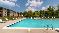 Located at 3901 Noyes Cir in Randallstown, Maryland, this outdoor pool facility serves as a central recreational amenity within the surrounding residential community. The centerpiece is a generously sized swimming pool with crystal-clear turquoise water, designed to accommodate a high volume of users. The pool is complemented by a broad, open concrete sun deck that lines multiple sides, offering ample space for lounging chairs, umbrella-shaded seating areas, and easy pool access via built-in entry steps and railings. Bordering one side of the pool area is a two-story brick building featuring bold red accent doors, upper-level balconies with black metal railings, and covered ground-floor access points that provide shade and shelter directly adjacent to the deck. The facility is enclosed by fencing and framed by mature trees and green landscaping, creating a sense of privacy and a natural backdrop that enhances the outdoor atmosphere. Additional multi-story residential brick buildings are visible in the background, reflecting the classic architectural character of the community. The open-sky setting, surrounded by lush foliage, provides a bright and welcoming environment well-suited for outdoor recreation and leisure throughout the warmer months.